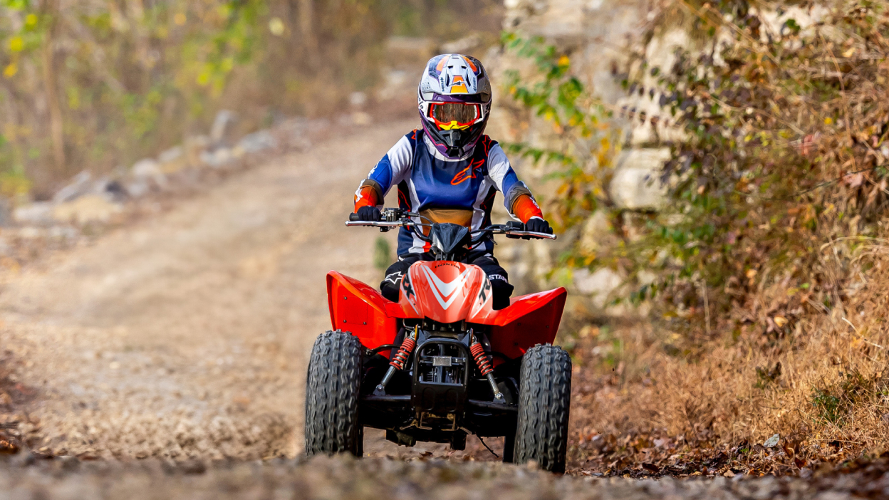 Young rider on ATV on dirt road. Grass and fence in background.