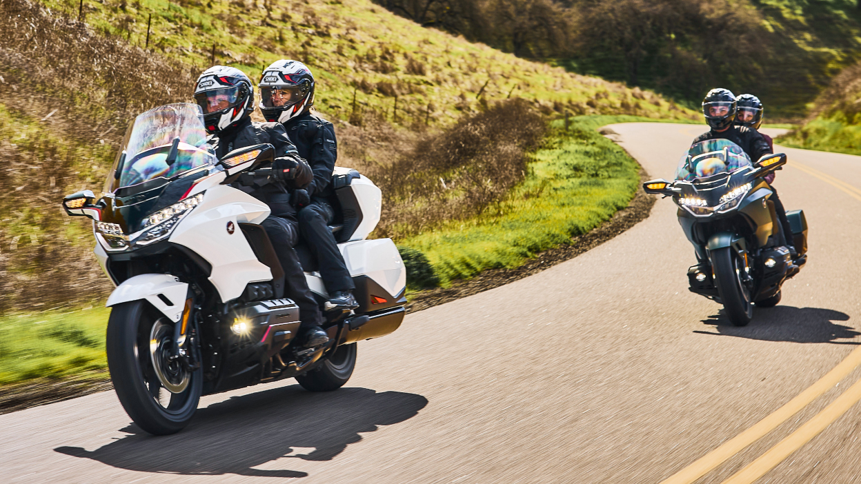Two riders on white bike infront of two riders on green bike on road. Hills in background.