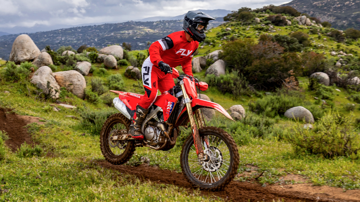 Rider going right on dirt track. Green field and hill with grey rocks in background.