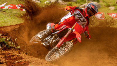 Rider front facing on motorbike on dirt track. Blue sky and green trees in background.
