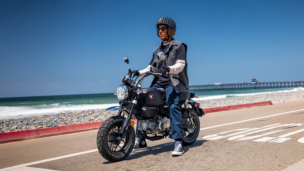 Rider on Motorbike. Stationary on road, facing left. Beach in background.