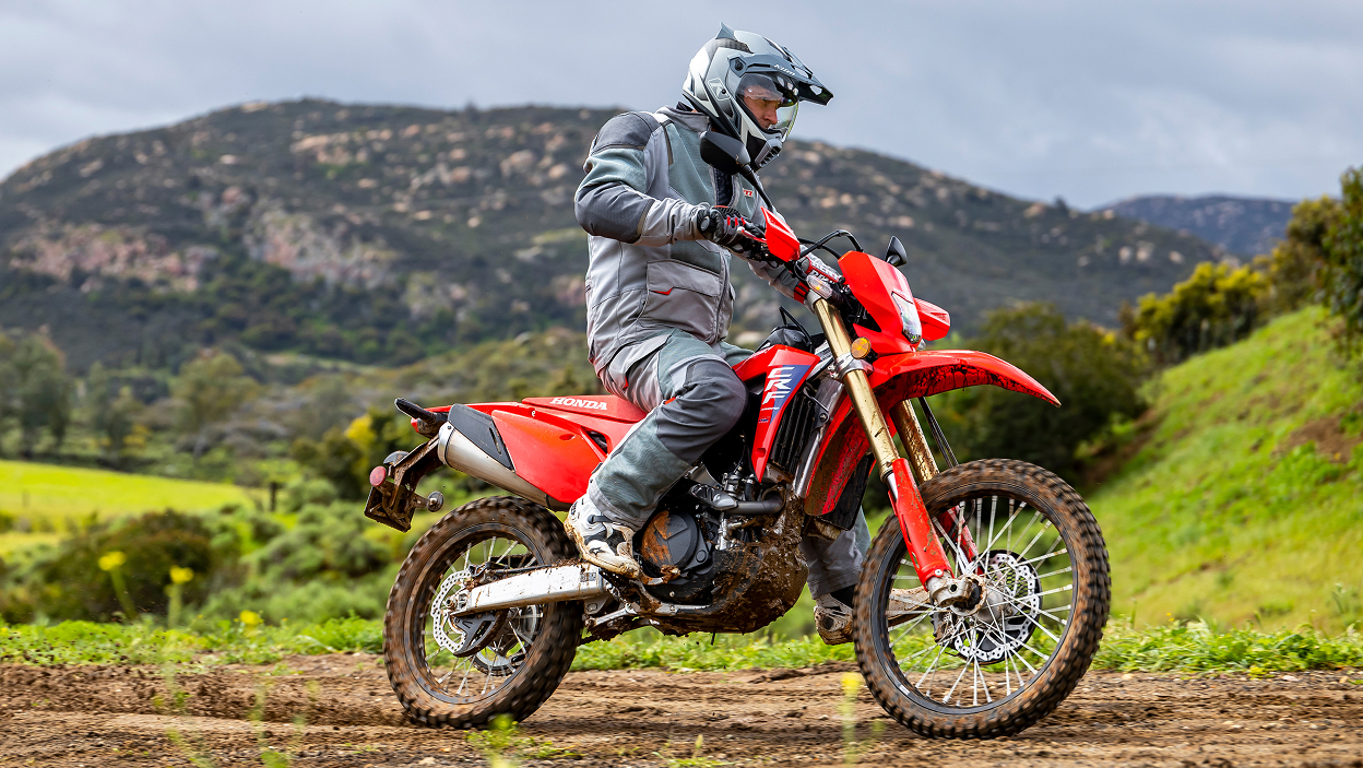 Rider on CRF450L on dirt road. Green bushes in background.